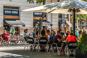 An outdoor seating area with tables and chairs under umbrellas, with a building featuring signage for 'CIUDAD COMTAL' in the background.