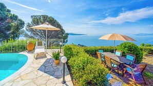 A poolside area with a table and chairs, surrounded by greenery and overlooking a body of water.