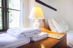 A neatly made bed with white linens, a bedside lamp, and a stack of folded towels on a wooden bedside table.