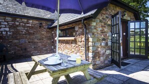 A stone building with a wooden table and chairs under a large umbrella, a stone wall, and a door with glass panels. - Delightful 2 bedroom cottage in beautiful Devon countryside, indoor pool & sauna (Tiverton)