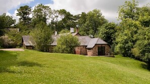 A stone house with a sloped roof, surrounded by a grassy lawn and trees. - Delightful 2 bedroom cottage in beautiful Devon countryside, indoor pool & sauna (Tiverton)