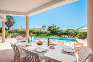 Une table à manger dressée pour quatre personnes, avec assiettes, couverts et une bouteille de vin, donnant sur une piscine et un jardin.
