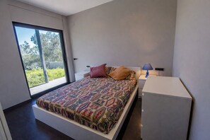A modern bedroom with a large bed, a bedside table with a lamp, and a view of greenery through a sliding door.