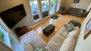 A spacious living room with a large flat-screen TV mounted on the wall, a grey sofa set, a black ottoman, and a dining area with a table and chairs.