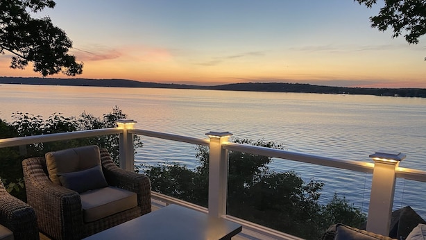A deck with wicker furniture and a table, overlooking a calm body of water at sunset.