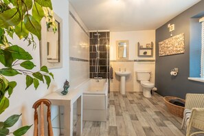 A bathroom with a bathtub, sink, toilet, and a shower. There are plants, a mirror, and a wall-mounted shelf. - 3 Bed in Appledore (oc-cockle) (Appledore)