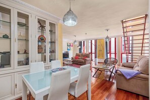 A spacious living area with a glass-top dining table, white chairs, a brown sofa, and a red-framed window. - Emma - Holiday Penthouse Malaga City (Málaga)