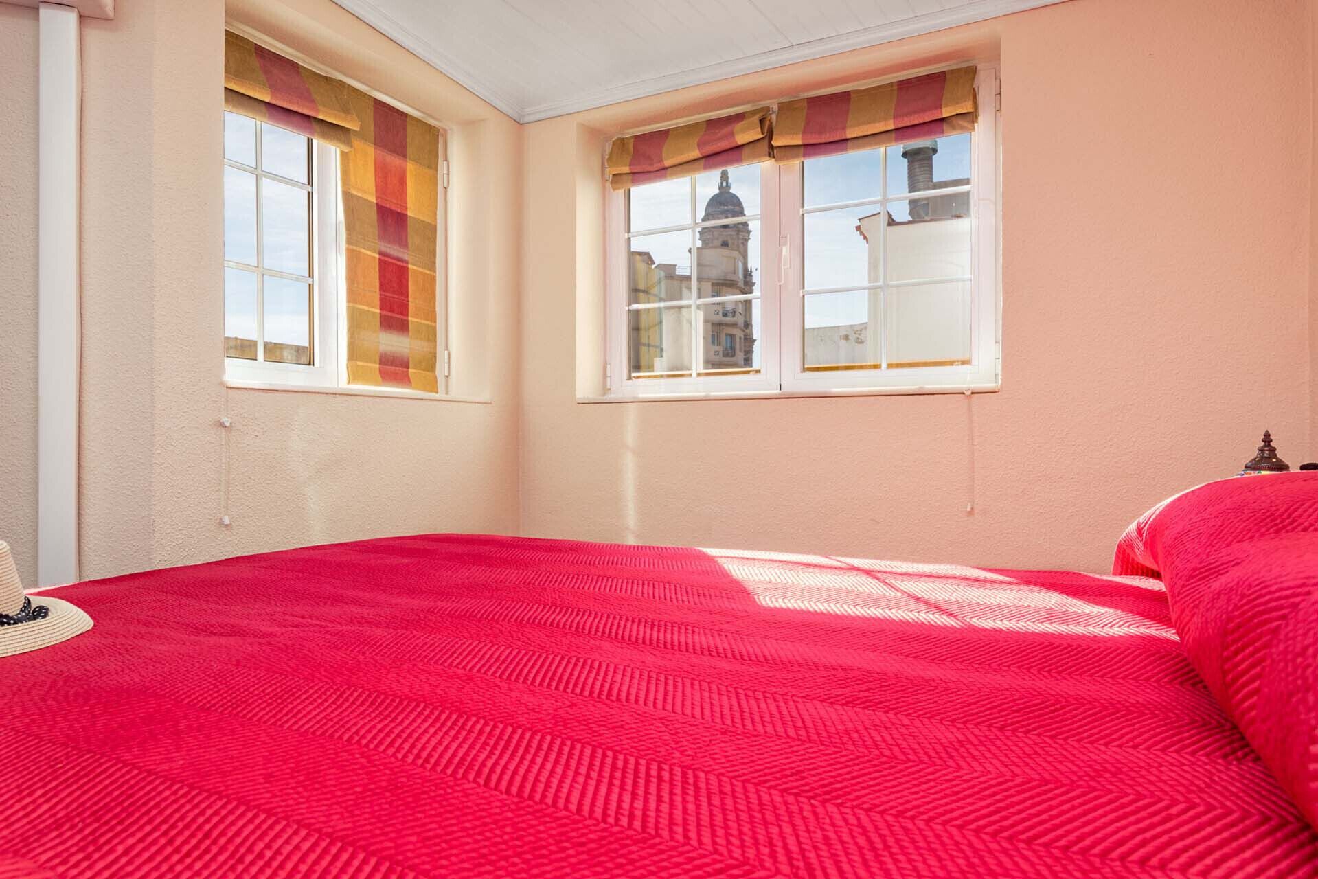 A bedroom with a red bedspread, two windows with curtains, and a view of a building outside.