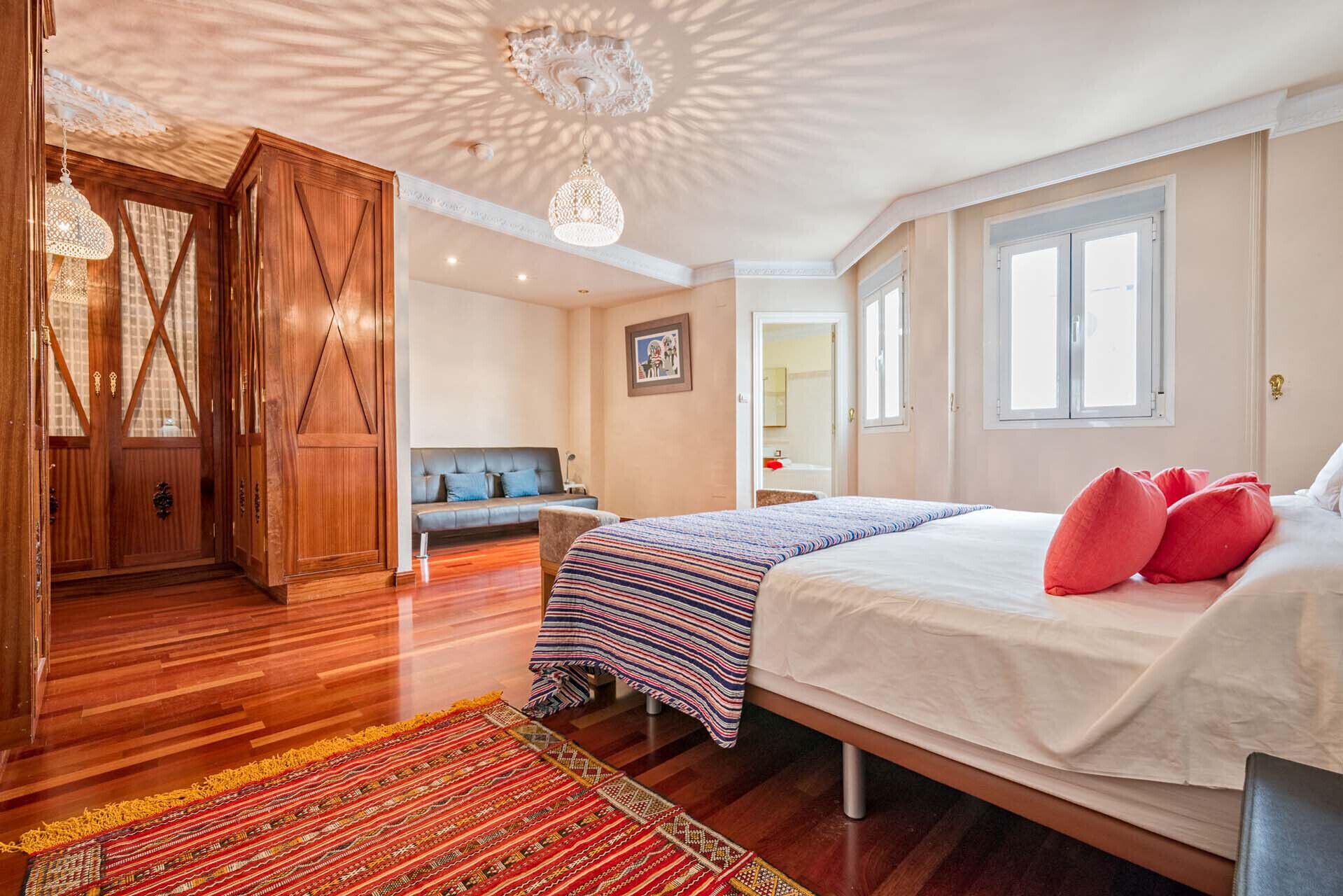 A bedroom with a bed, a sofa, a wooden wardrobe, and a patterned rug.
