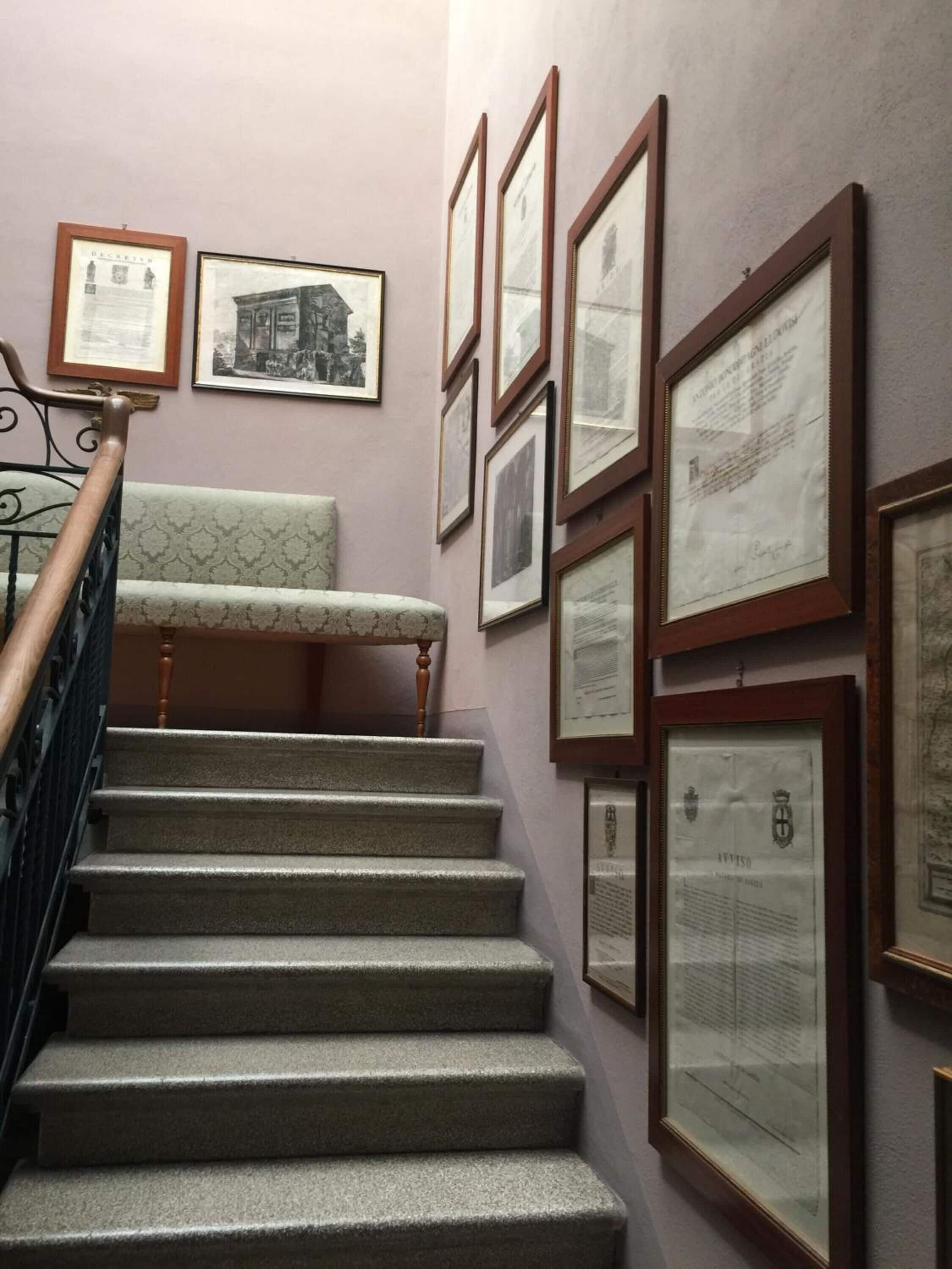 Un escalier recouvert d’une moquette à motifs et doté d’une rampe en bois, menant à une pièce dont le mur est également décoré de motifs et où sont accrochés plusieurs documents ou certificats encadrés.
