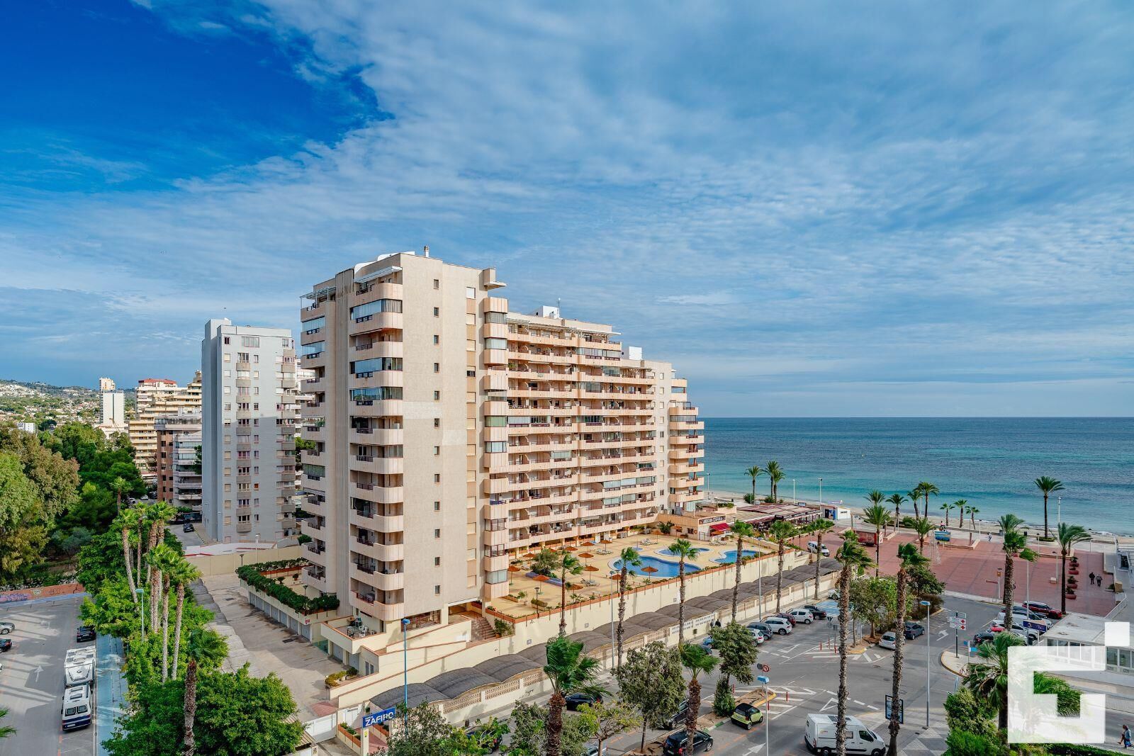 A multi-story hotel with balconies, a swimming pool, and a view of the sea.