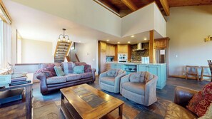 Amplia sala de estar con sofá, sillones, una mesa de centro de madera y una cocina al fondo.