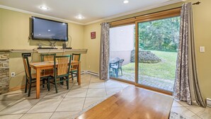 A room with a wooden dining table and chairs, a flat-screen TV mounted on the wall, and a sliding glass door leading to a patio with a stone wall and greenery.