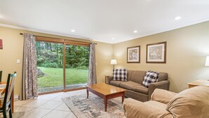 A living room with a sofa, coffee table, and a dining area with chairs. There is a sliding glass door leading to a garden.