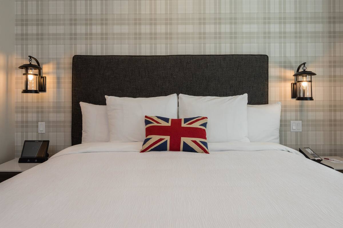 Un lit bien fait, avec du linge de lit blanc et un oreiller orné du motif Union Jack. Deux tables de chevet sont fixées au mur, chacune étant surmontée d’un luminaire de style lanterne noir. Une tablette est posée sur l’une des tables de chevet.