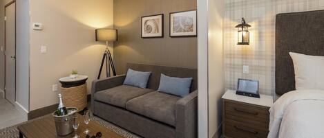 Habitación de hotel con un sofá gris, una mesita de noche con lámpara, una mesita de noche más pequeña con un reloj digital y una alfombra estampada.