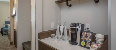 A hotel room with a kitchenette area featuring a coffee maker, glassware, and a coffee pod holder.