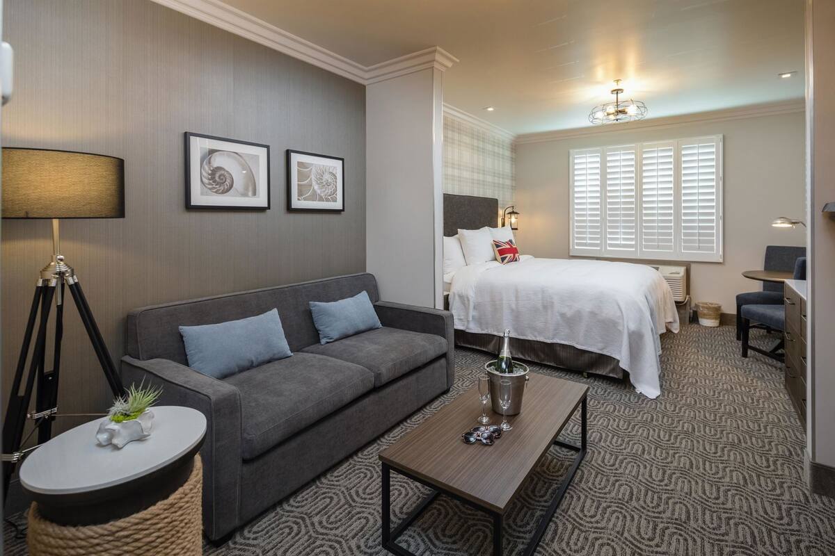 Une chambre d’hôtel avec un canapé gris, un lit avec une literie blanche, une petite table sur laquelle se trouvent une bouteille et des verres, et une lampe.