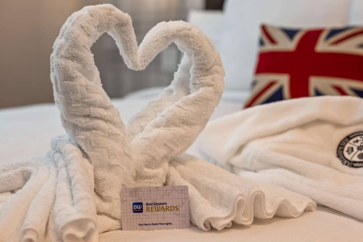 Une serviette blanche, pliée avec soin et en forme de cœur, et une autre serviette ornée d’un logo circulaire, sont posées sur un lit.