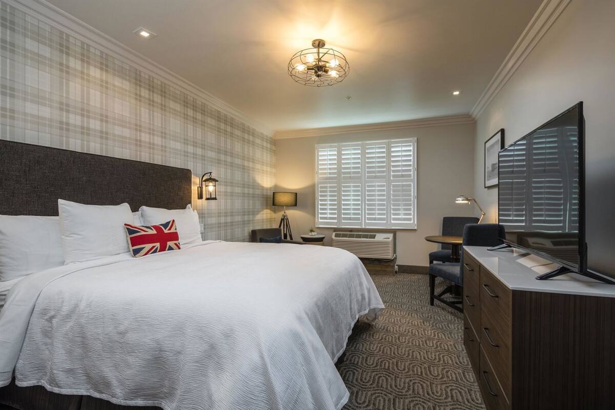 Une chambre d’hôtel avec un grand lit, un bureau, une chaise, une télévision et une fenêtre avec des stores.