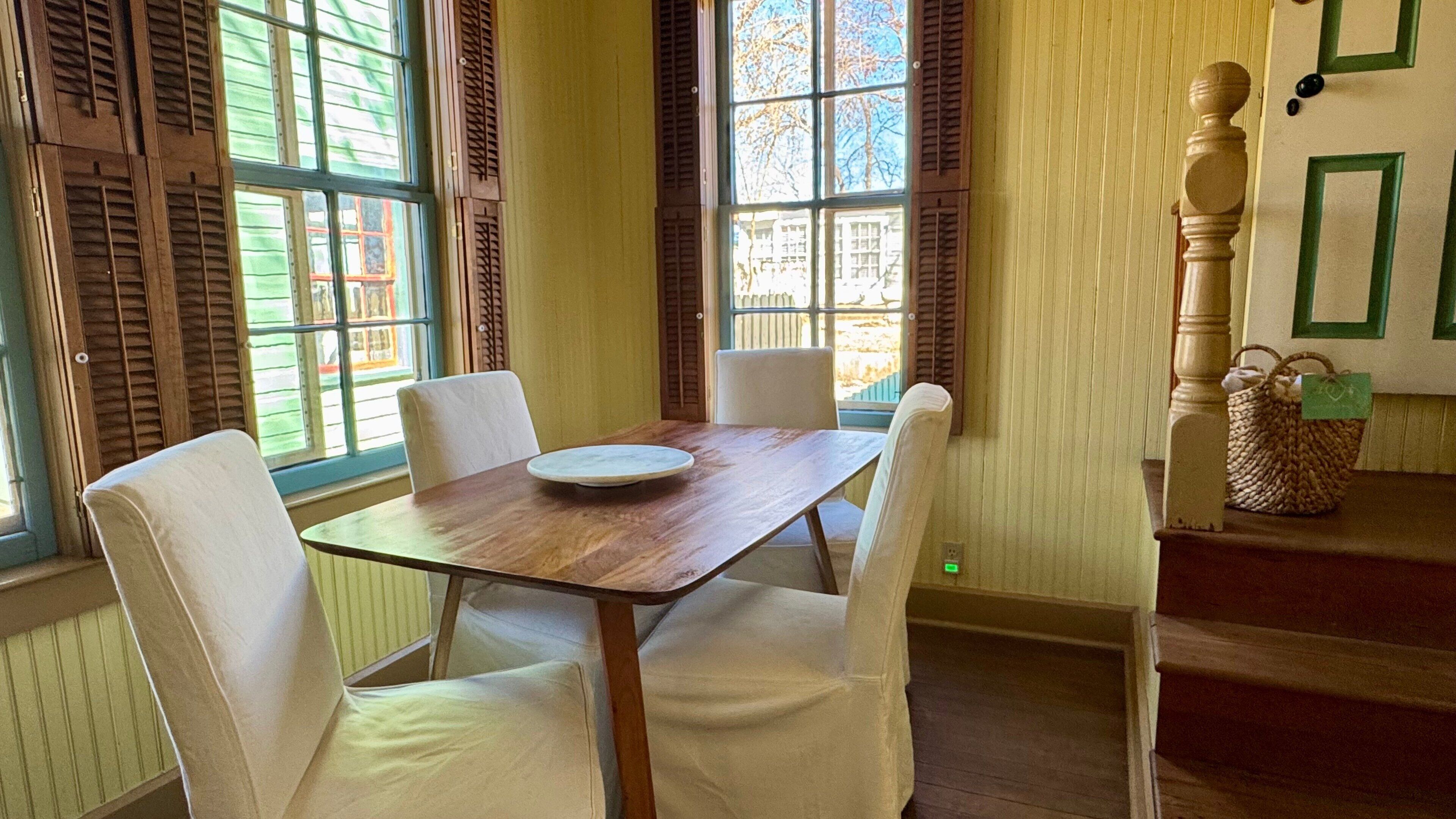 Un coin repas avec une table en bois et quatre chaises blanches, de grandes fenêtres avec des volets et un escalier orné d’un panier sur la plateforme.