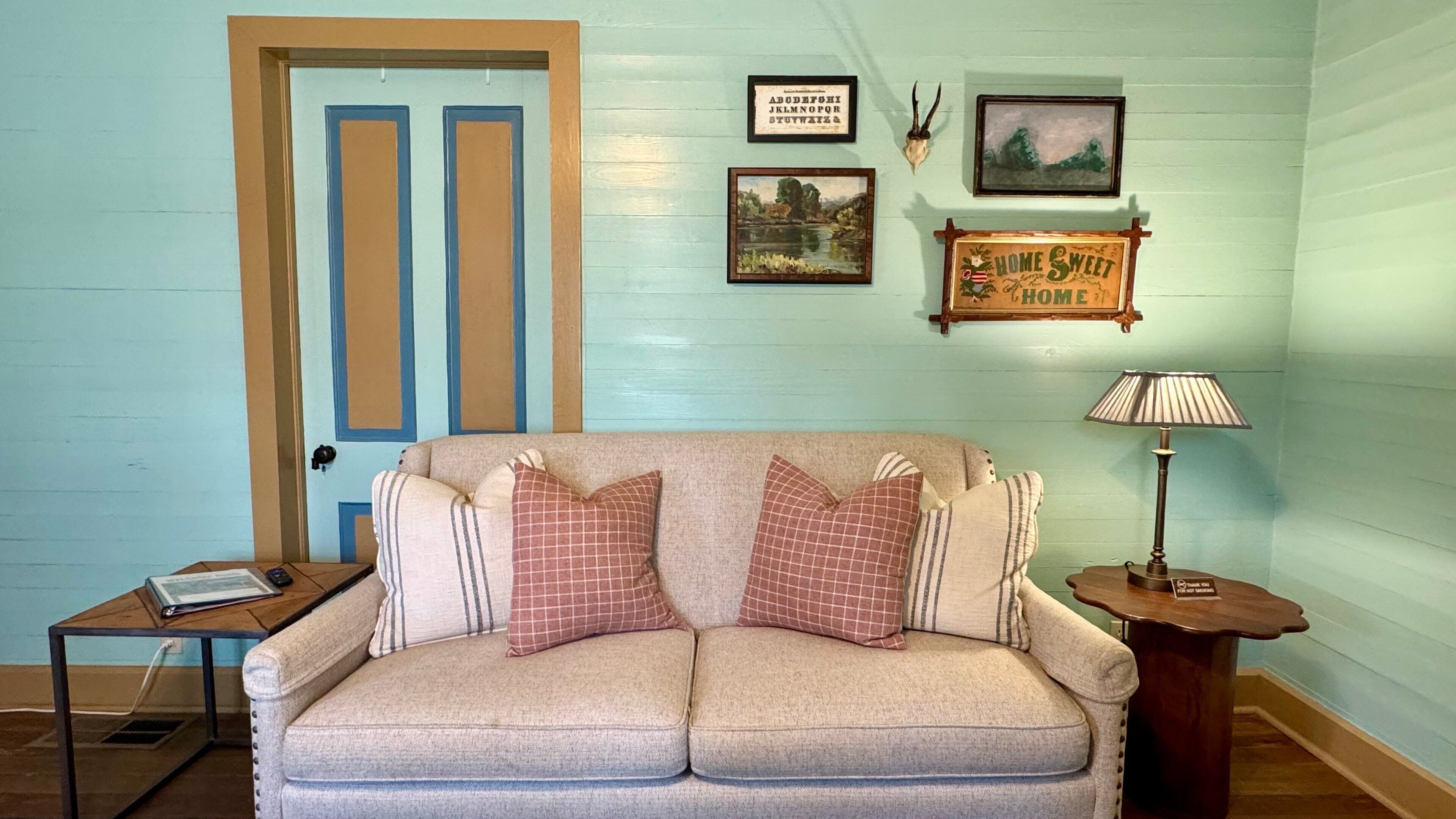 Une pièce chaleureuse avec un mur bleu clair, un canapé beige agrémenté de coussins à carreaux, une petite table sur laquelle se trouve un livre, une lampe, et des tableaux encadrés accrochés au mur.