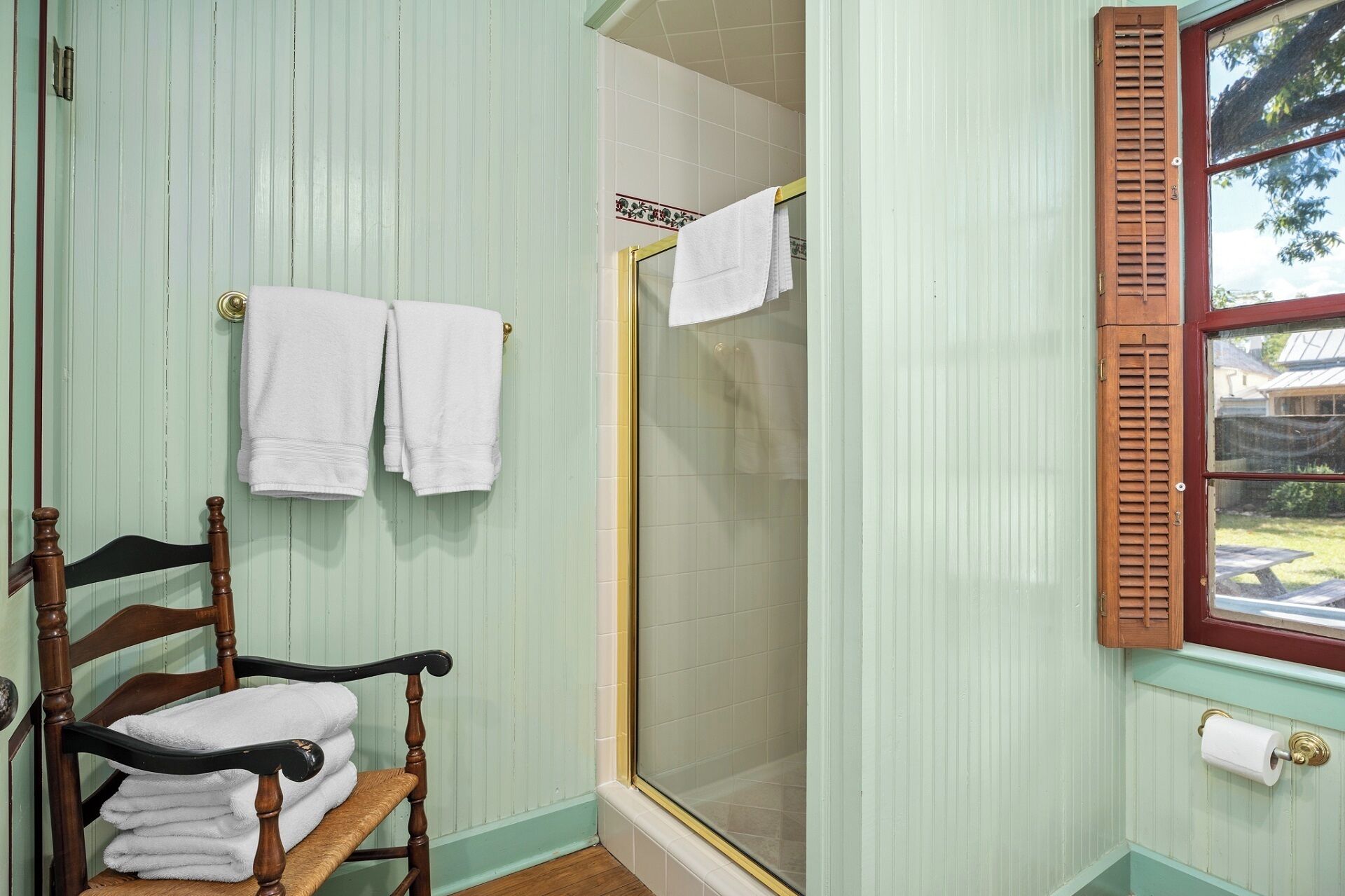Une salle de bain avec une chaise en bois, deux serviettes accrochées au mur, une porte de douche en verre et une fenêtre avec des volets en bois.