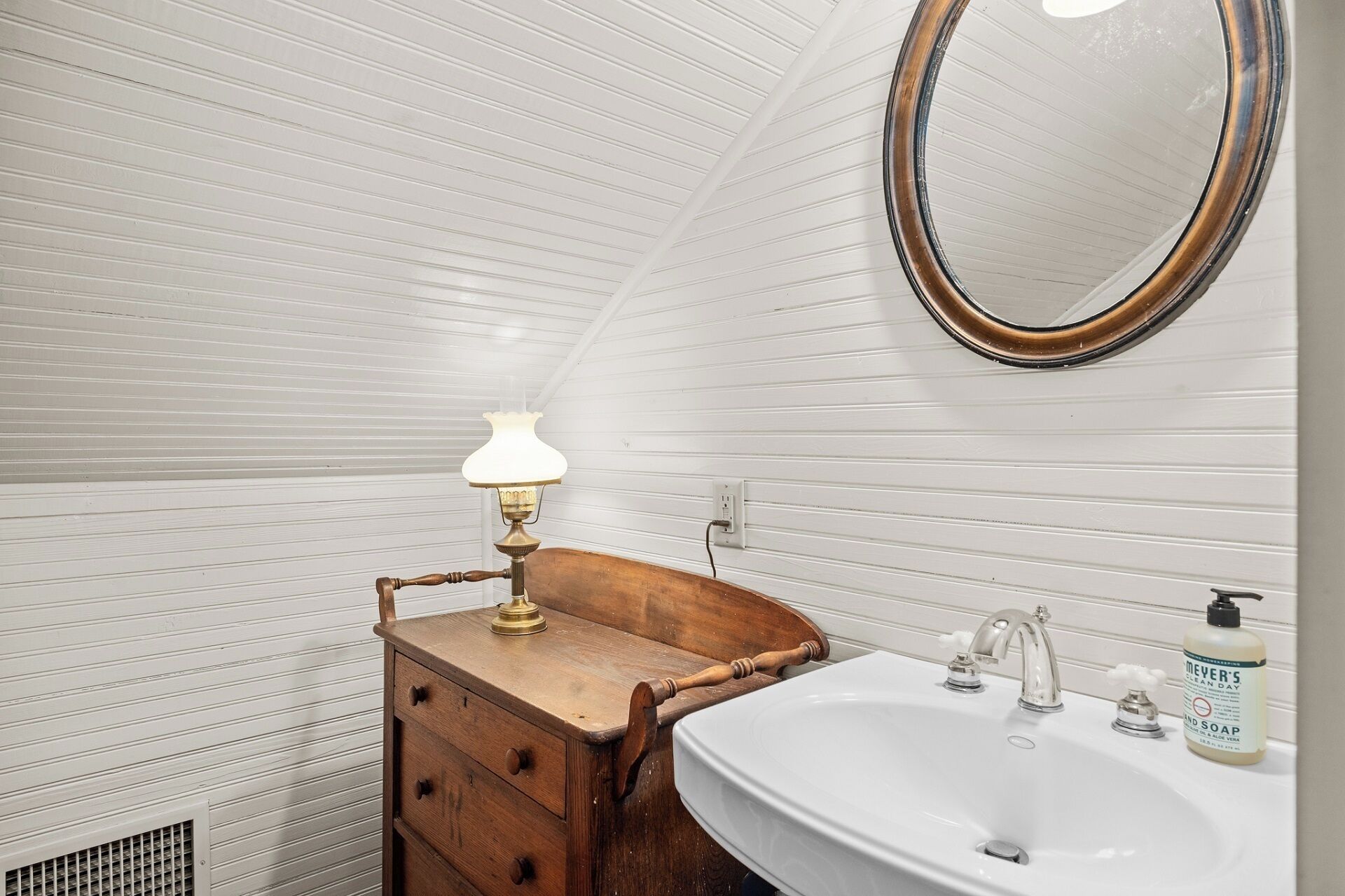 A compact bathroom with a wooden vanity, a white sink, a mirror, and a wall-mounted lamp.
