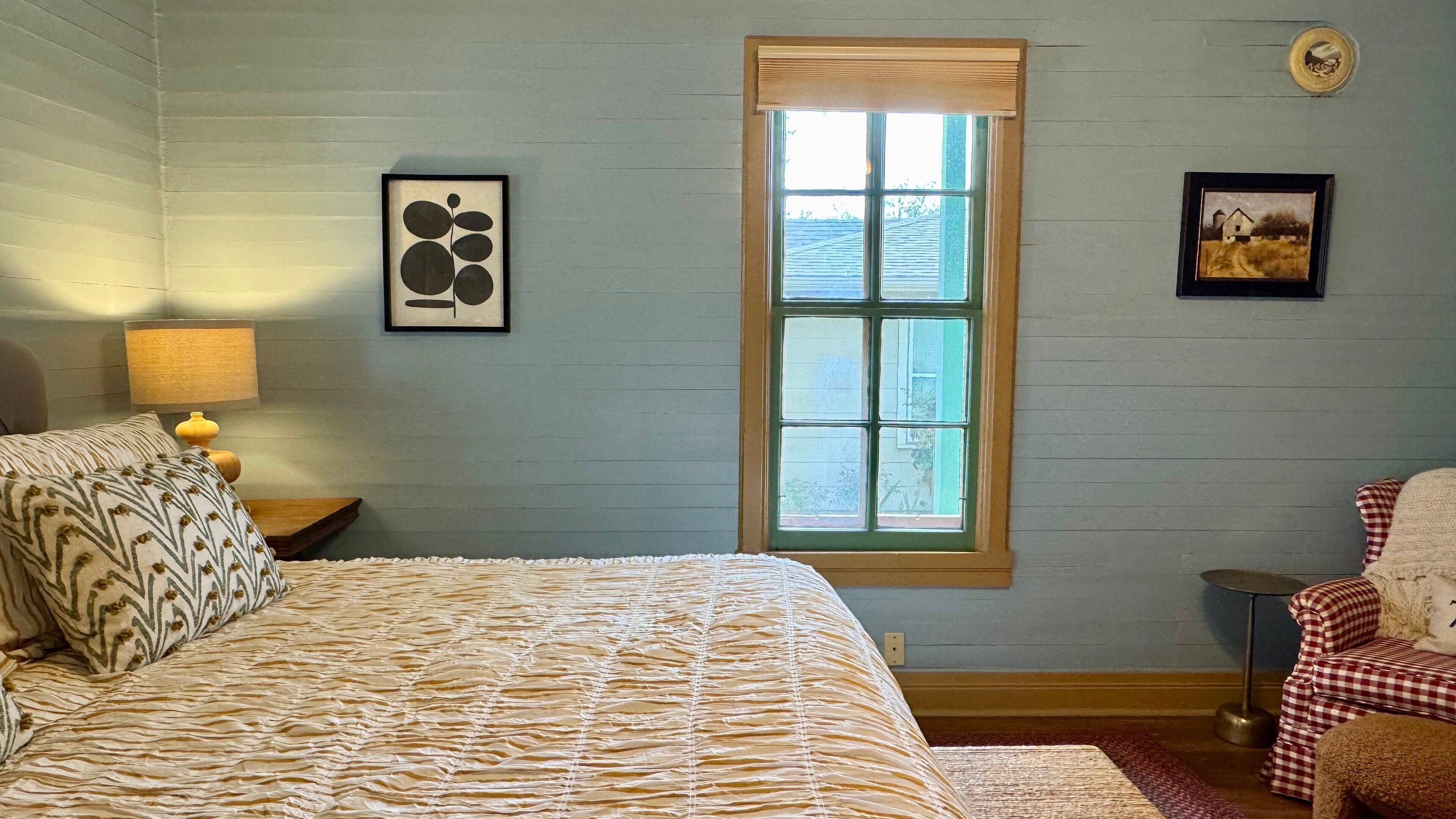 A bedroom with a large bed, a window, a lamp, and two framed pictures on the wall.