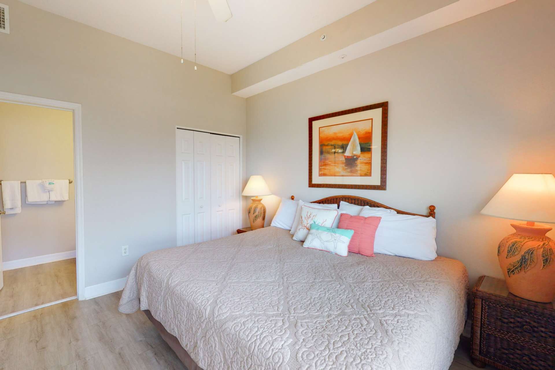 A bedroom with a large bed, two bedside lamps, a door with a towel rack, and a painting on the wall.