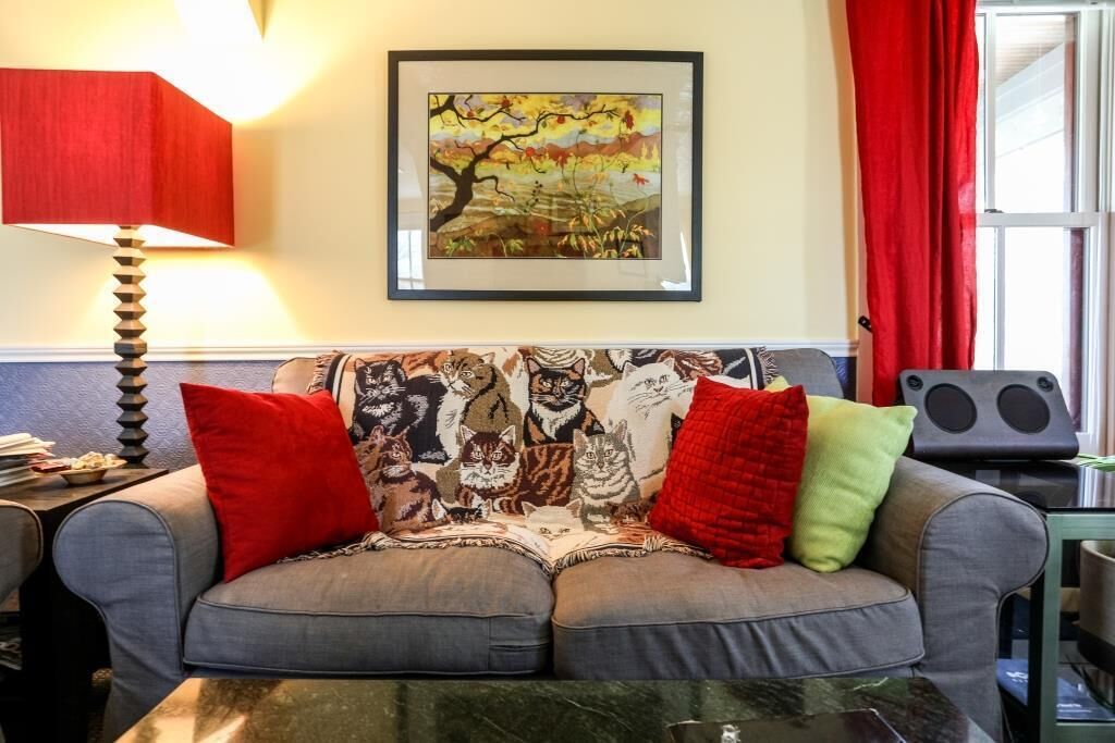 Una sala de estar con un sofá decorado con un cojín con motivos de gato, un cuadro en la pared y una lámpara roja.