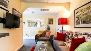 A living room with a sofa, a coffee table, a TV, and a red lamp. - Cyclismo - Suite 2 | Close to Main St. | Wonderful Porch (Fredricksburg)