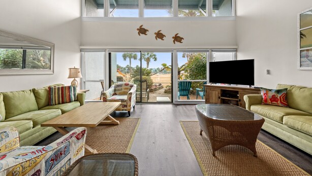A spacious living room with a large TV, green sofas, a wicker table, and a view of palm trees through the glass doors. - Unit 27, The Sail Away, 2 Bedroom Beach Condo (Panama City Beach)