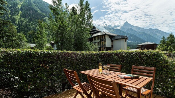 Una terrazza in legno con tavolo e sedie, affacciata su un paesaggio montuoso con un edificio sullo sfondo.