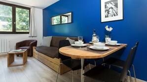 Una stanza moderna con una parete azzurra, un tavolo da pranzo in legno apparecchiato con piatti e bicchieri, un divano marrone e una finestra con vista sugli alberi.