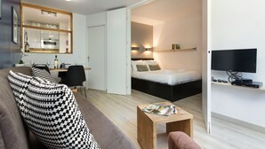 Una camera d'albergo moderna con un divano, un piccolo tavolino, un letto, una TV e una zona cucina visibile attraverso una porta aperta.