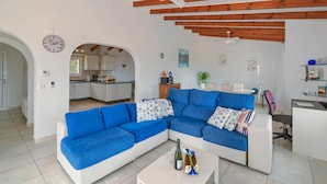 Uma sala de estar moderna com um sofá seccional azul, uma mesa de centro branca com bebidas e uma cozinha ao fundo.