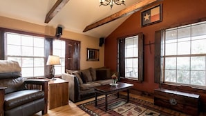 Una sala de estar acogedora con un sofá de cuero, una mesa de centro de madera y un tapete estampado. Hay ventanas grandes con marcos de madera y una decoración en forma de estrella en la pared.