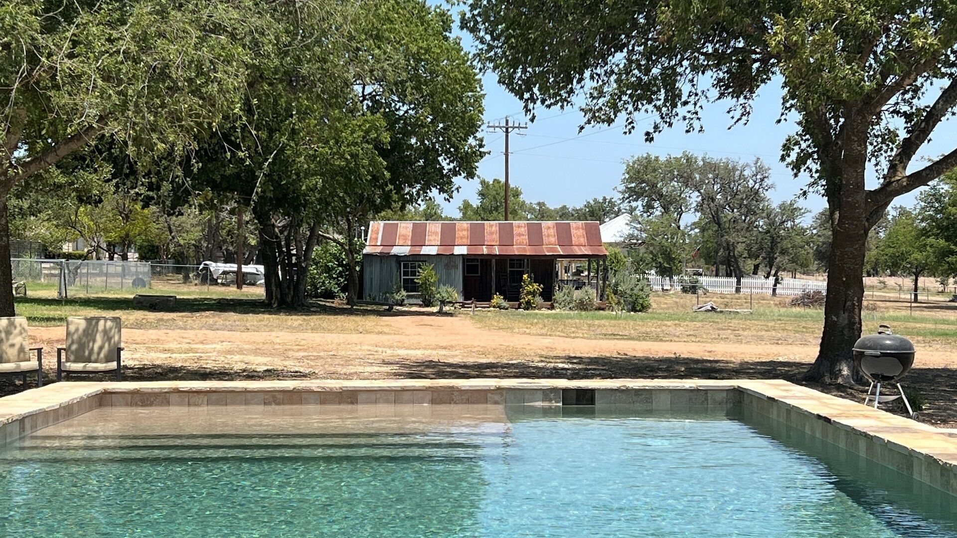 Une piscine avec une terrasse en bois, un bâtiment rustique doté d’un toit en métal, et des arbres en arrière-plan.