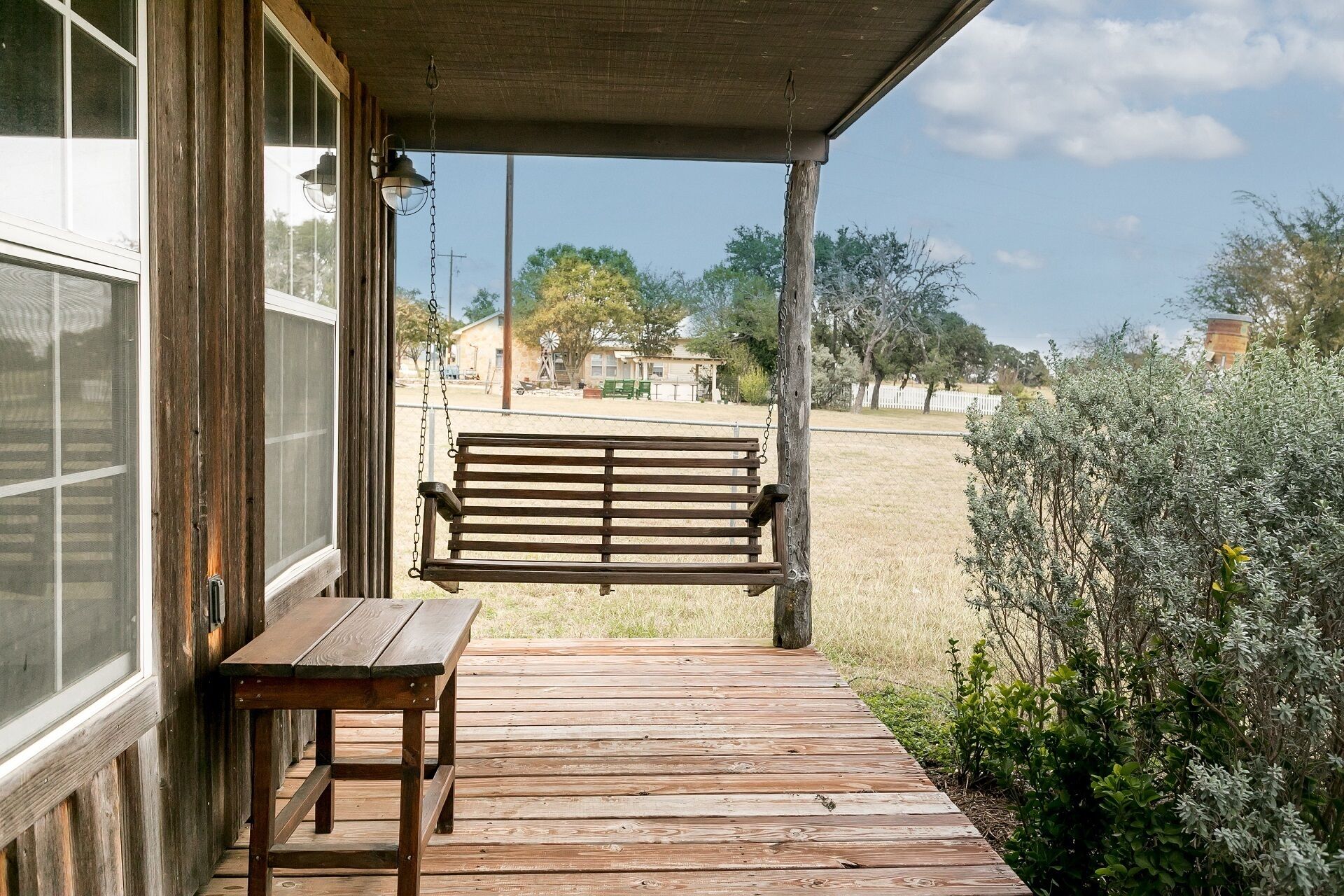 Une véranda en bois avec un banc et une table, donnant sur un espace verdoyant avec des arbres et un bâtiment situé au loin.