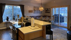 Una cocina amplia con una isla central, un comedor con sillas, un ventanal con vistas a árboles y un ventilador de techo.