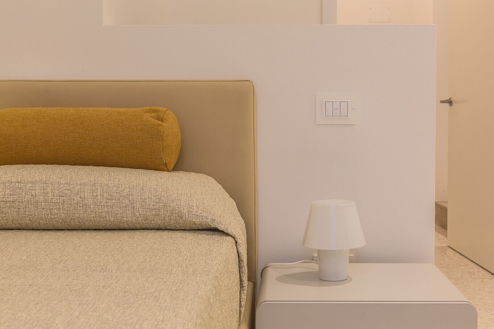 A neatly made bed with a textured blanket and a single pillow, a bedside table with a lamp, and a wall-mounted light switch.