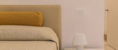 A neatly made bed with a textured blanket and a single pillow, a bedside table with a lamp, and a wall-mounted light switch.