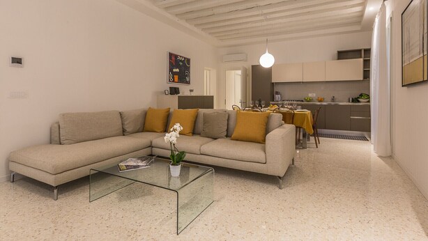A modern living room with a sectional sofa, a glass coffee table, and a dining area with a kitchen in the background. - Ca' Rossini 1 Apartment (Venice)