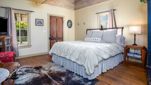 A bedroom with a bed, a wooden floor, a ceiling fan, a window with curtains, a door, a small table with a lamp, and a television on the left. - Das Solheid Sunday House | Close to Main (Fredricksburg)