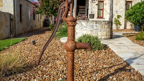 A rustic outdoor setting with a vintage water pump, stone buildings, and a well-maintained garden. - Das Solheid Sunday House | Close to Main (Fredricksburg)