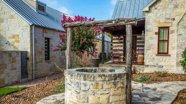 A stone well, a wooden pergola with pink flowers, and a stone building with a window and door. - Das Solheid Sunday House | Close to Main (Fredricksburg)