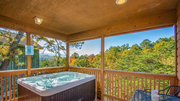 A wooden deck with a hot tub, a table with two cups, and a view of trees and mountains. - Ladybug Resort Romantic Cabin with Mountain Views (Sevierville)