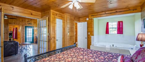 A wooden cabin bedroom with a bed, a ceiling fan, a bathroom with a bathtub, and a window with curtains.
