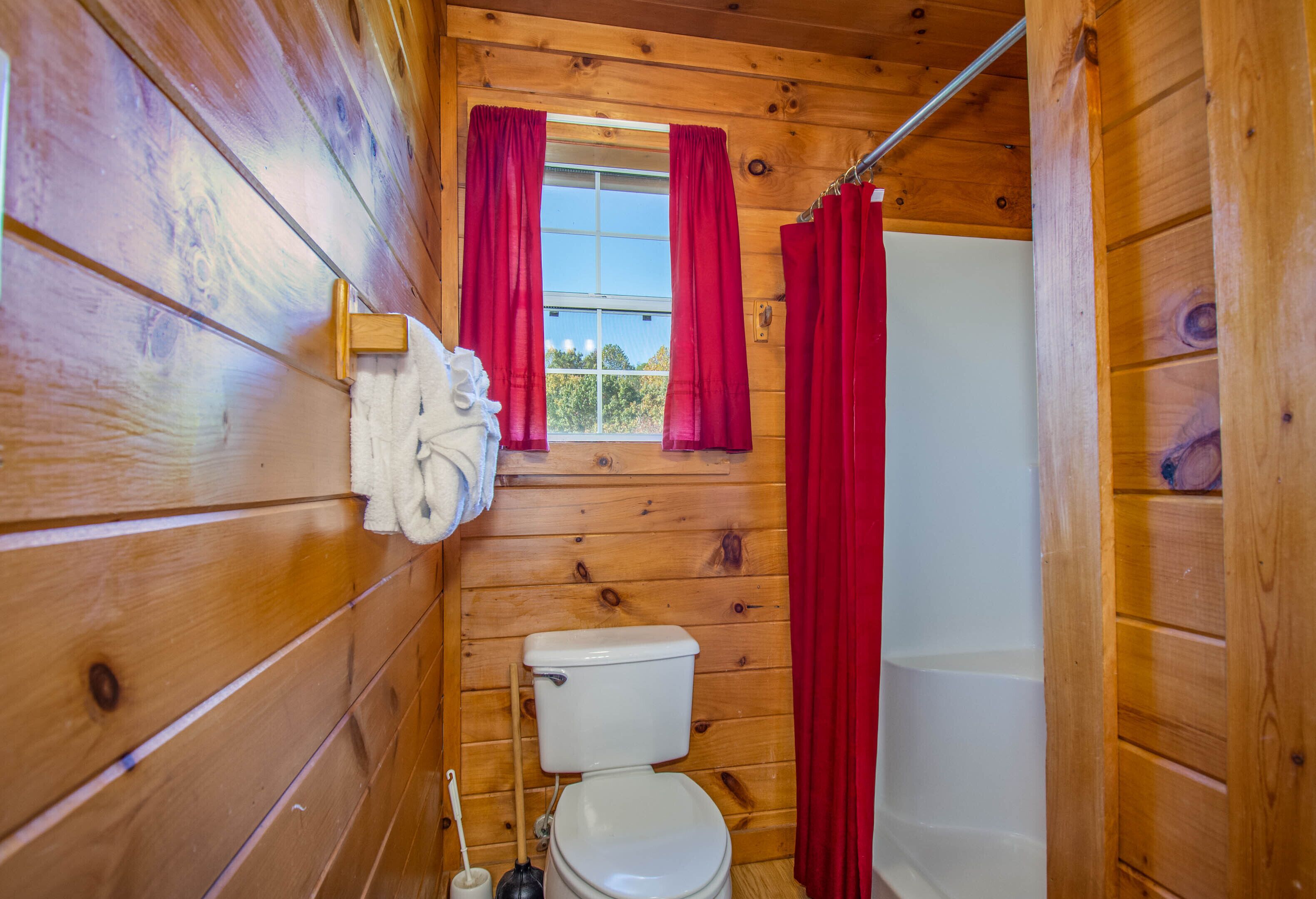 A wooden-paneled bathroom with a toilet, a window with red curtains, and a shower area.